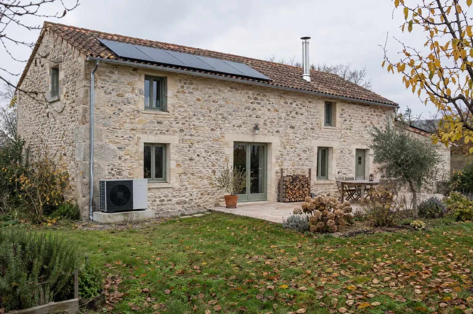 Maison en pierre rénovée avec unité extérieure de pompe à chaleur, panneaux solaires sur le toit, conduit de poêle et réserve de bois